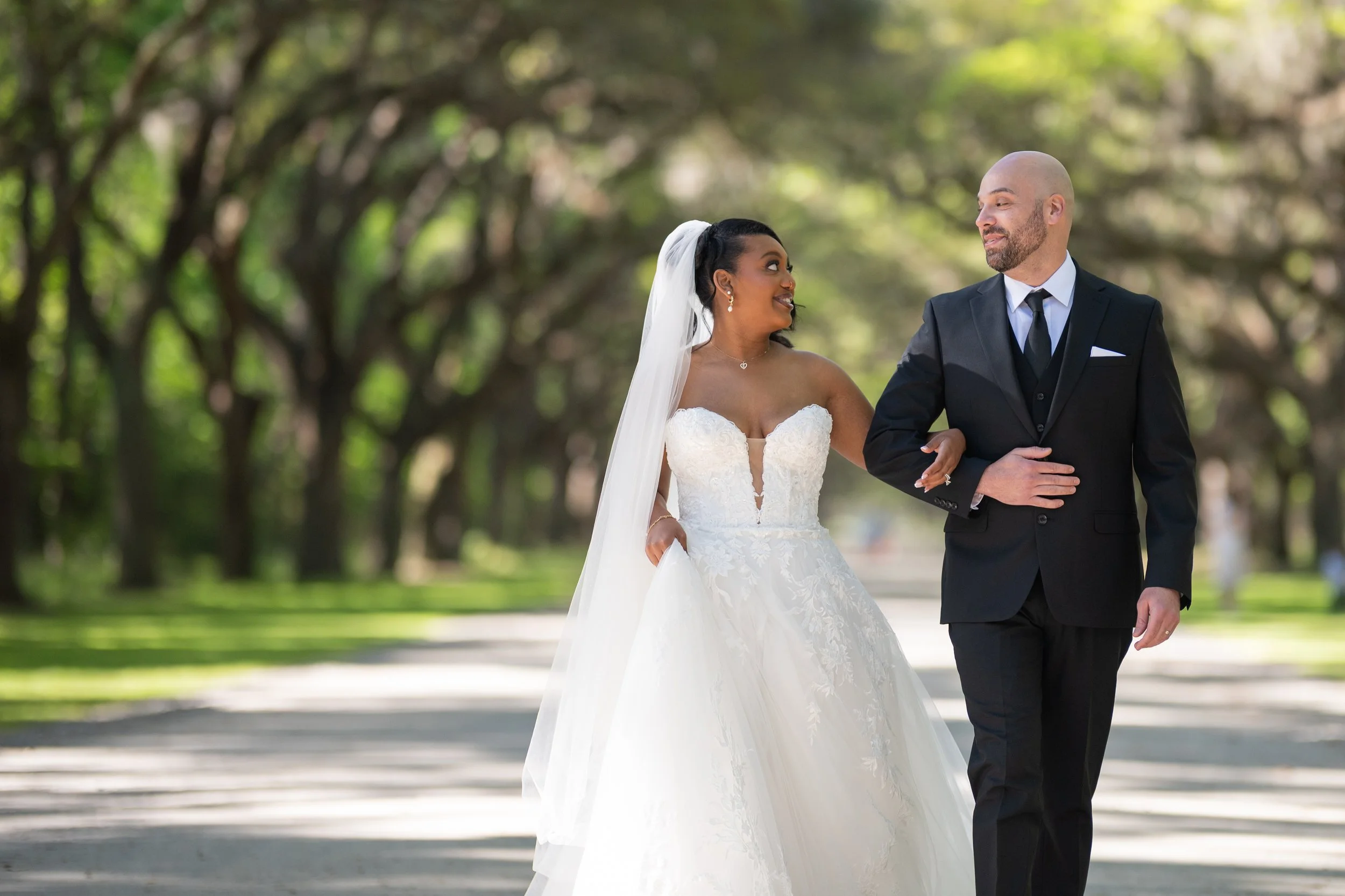 A bride and groom walking arm in arm outdoors, surrounded by green trees, on their wedding day.