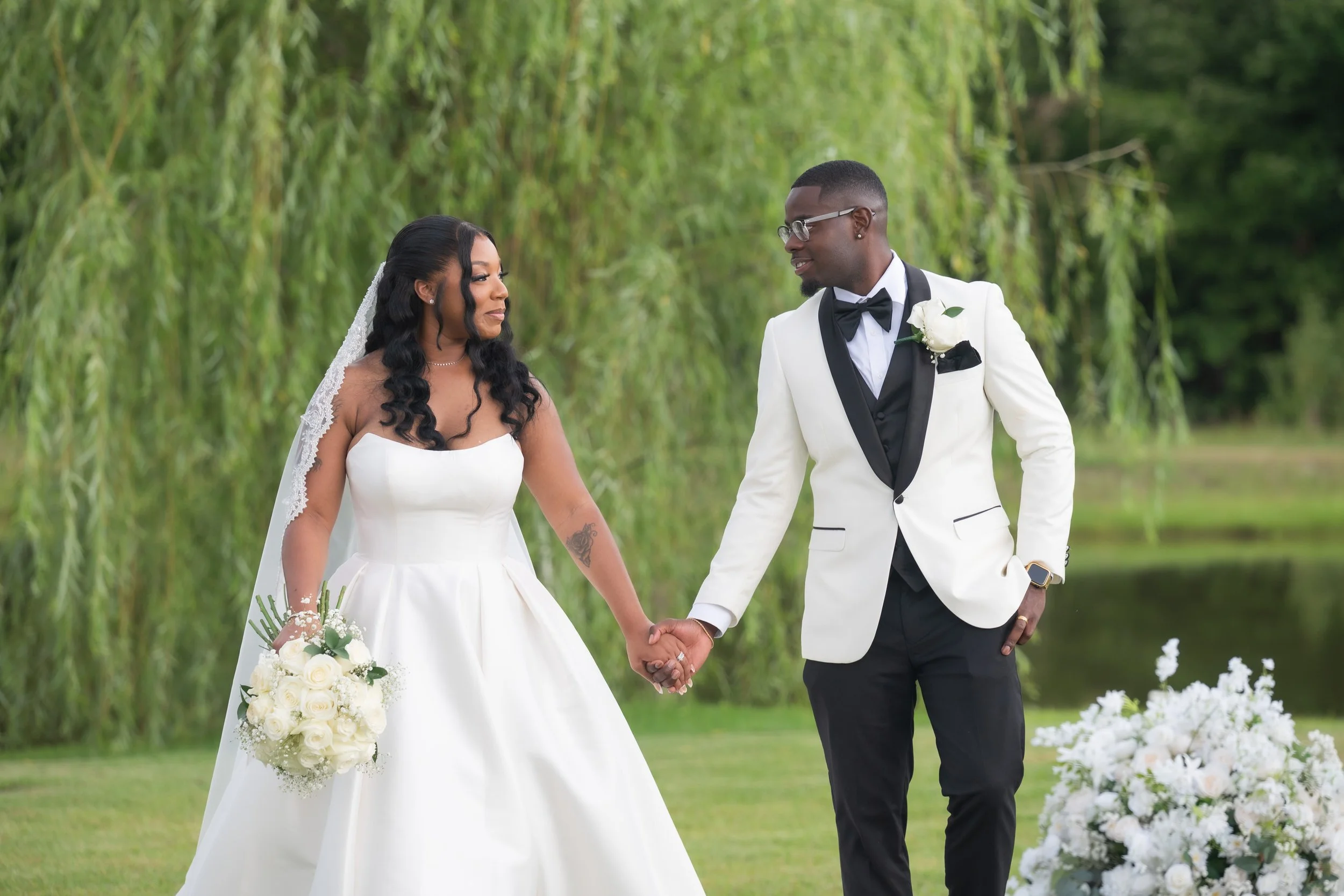 A bride and groom holding hands outdoors, with the bride in a white wedding gown and the groom in a white tuxedo jacket with black lapels and a black bow tie, standing near a body of water and surrounded by trees and greenery.