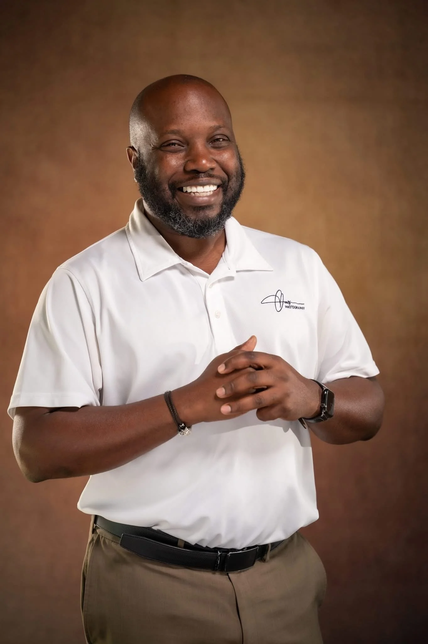 A smiling man with a beard, wearing a white polo shirt and khaki pants, standing with his hands clasped, against a brown background.