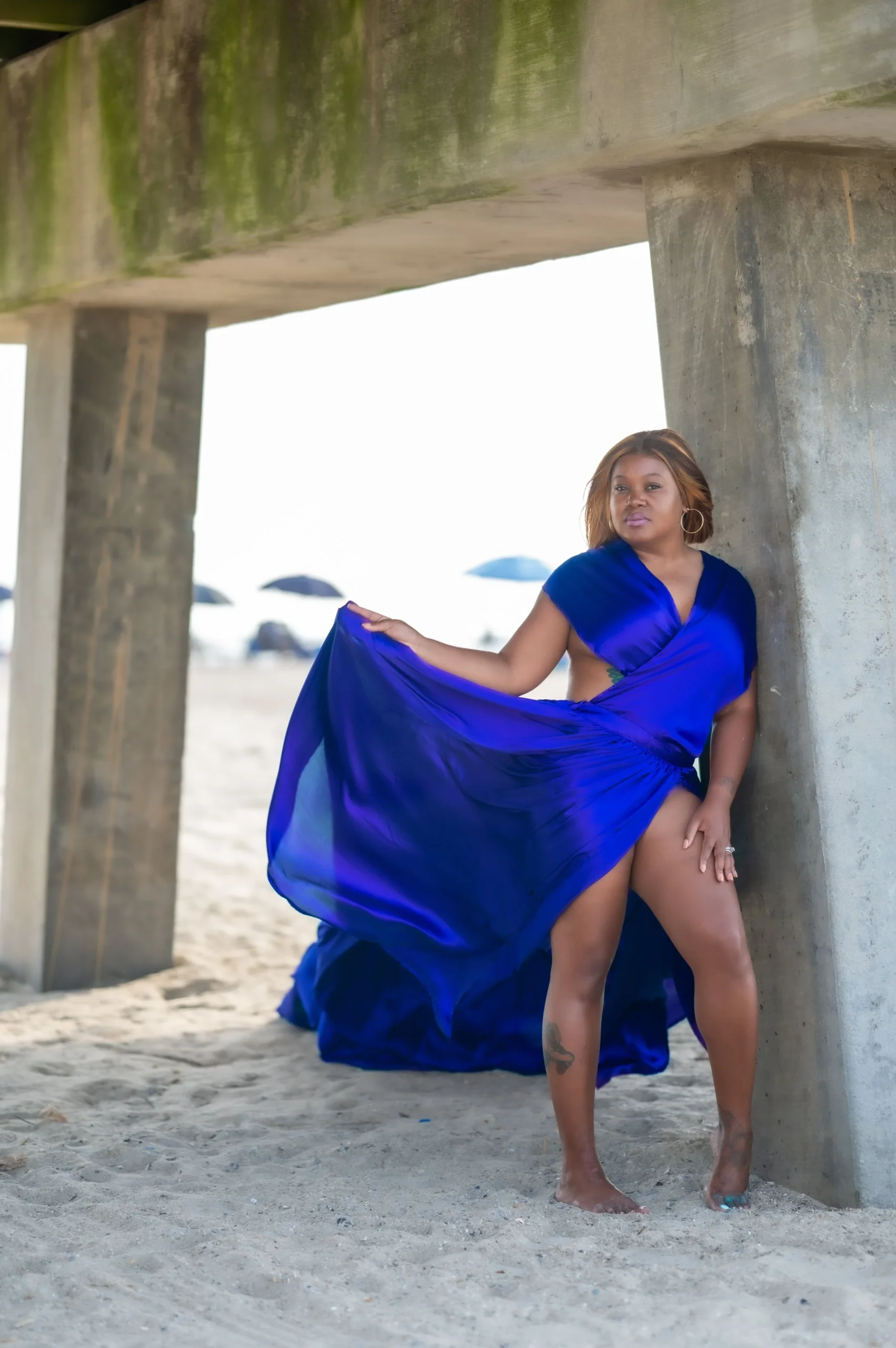 A woman in a flowing, vibrant blue dress standing under a concrete bridge on the beach with umbrellas in the background.