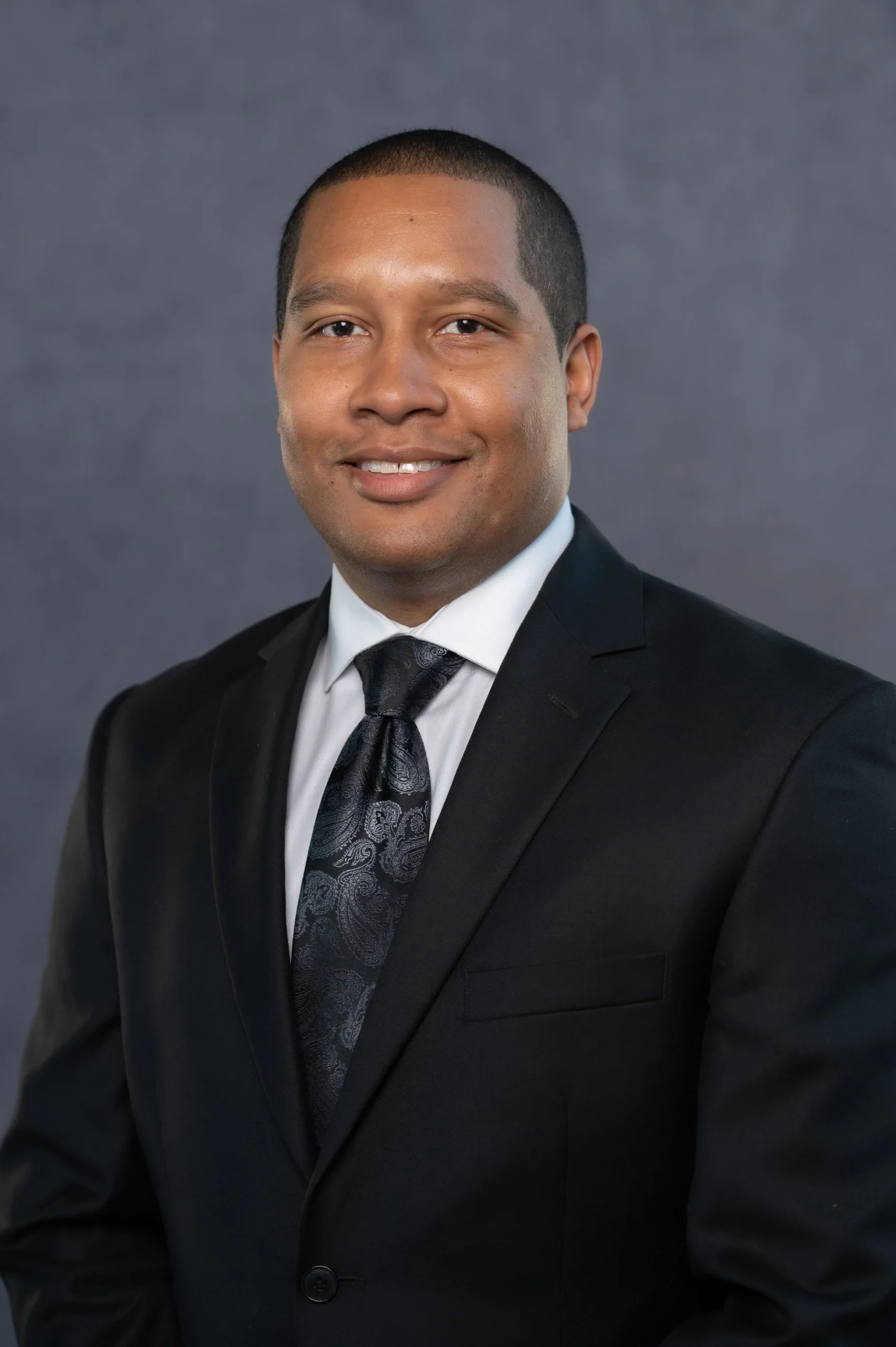 Professional portrait of a man with short black hair wearing a black suit, white shirt, and a patterned black tie against a gray background.