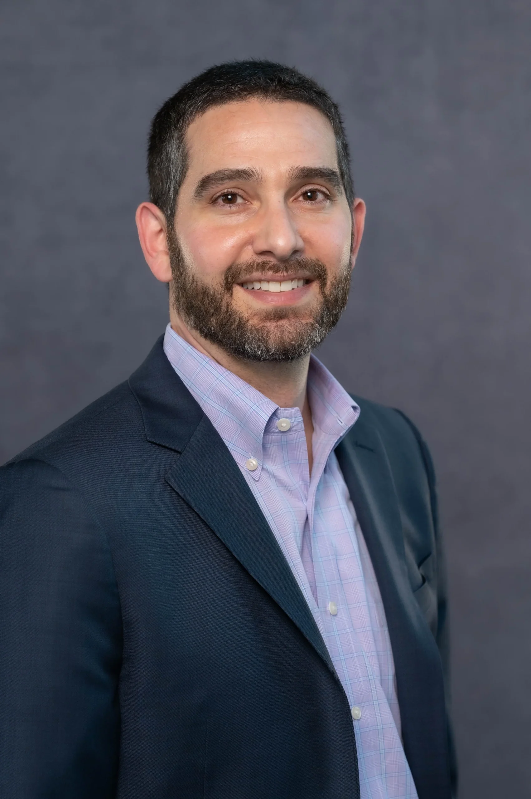 A man with dark hair, a trimmed beard, wearing a dark suit jacket and a light purple checkered dress shirt, smiling at the camera in a professional portrait.