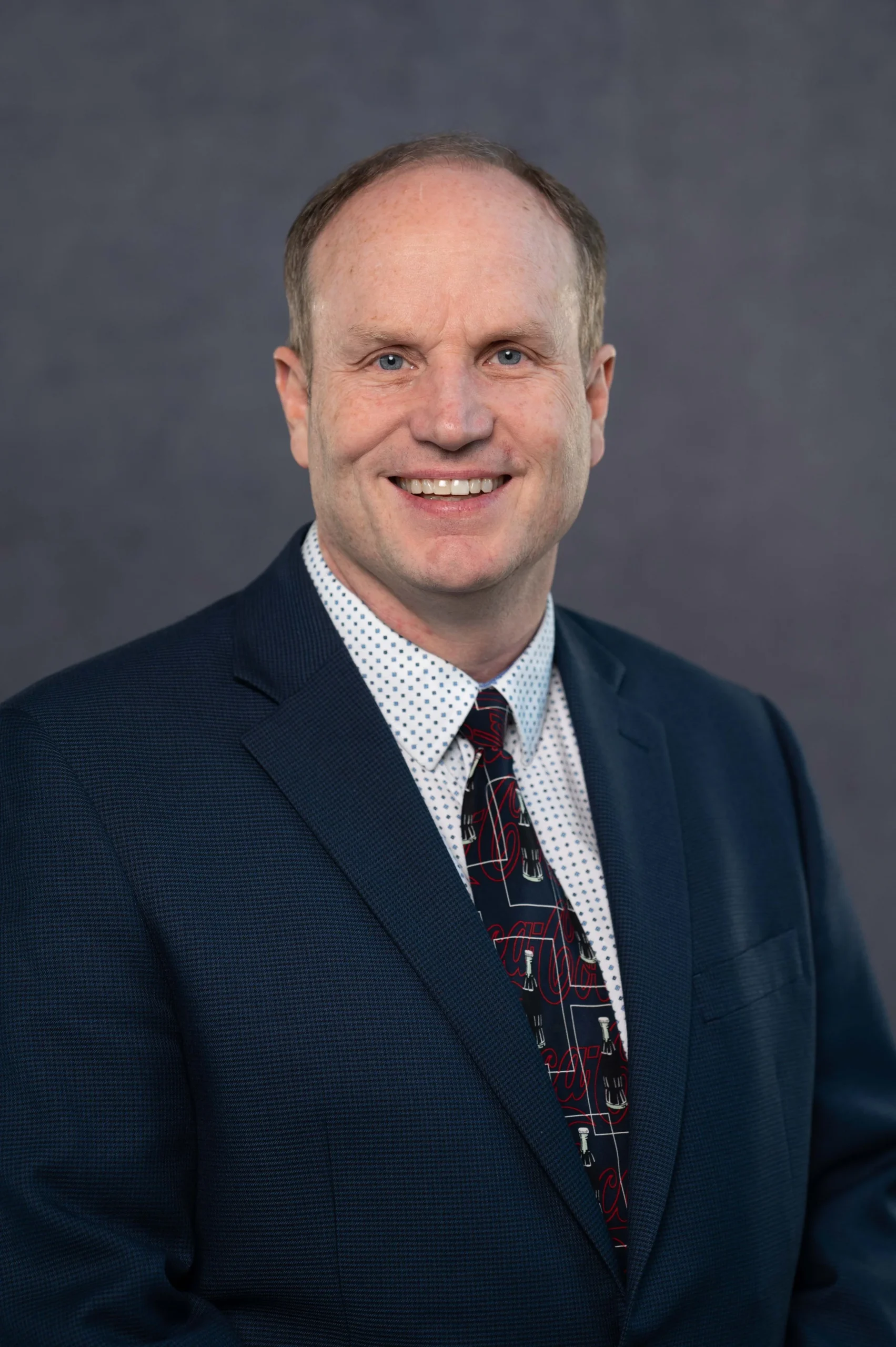 A smiling man with short light brown hair, wearing a dark blue suit, white dress shirt with small blue dots, and a black tie with red and white designs, posing against a gray background.
