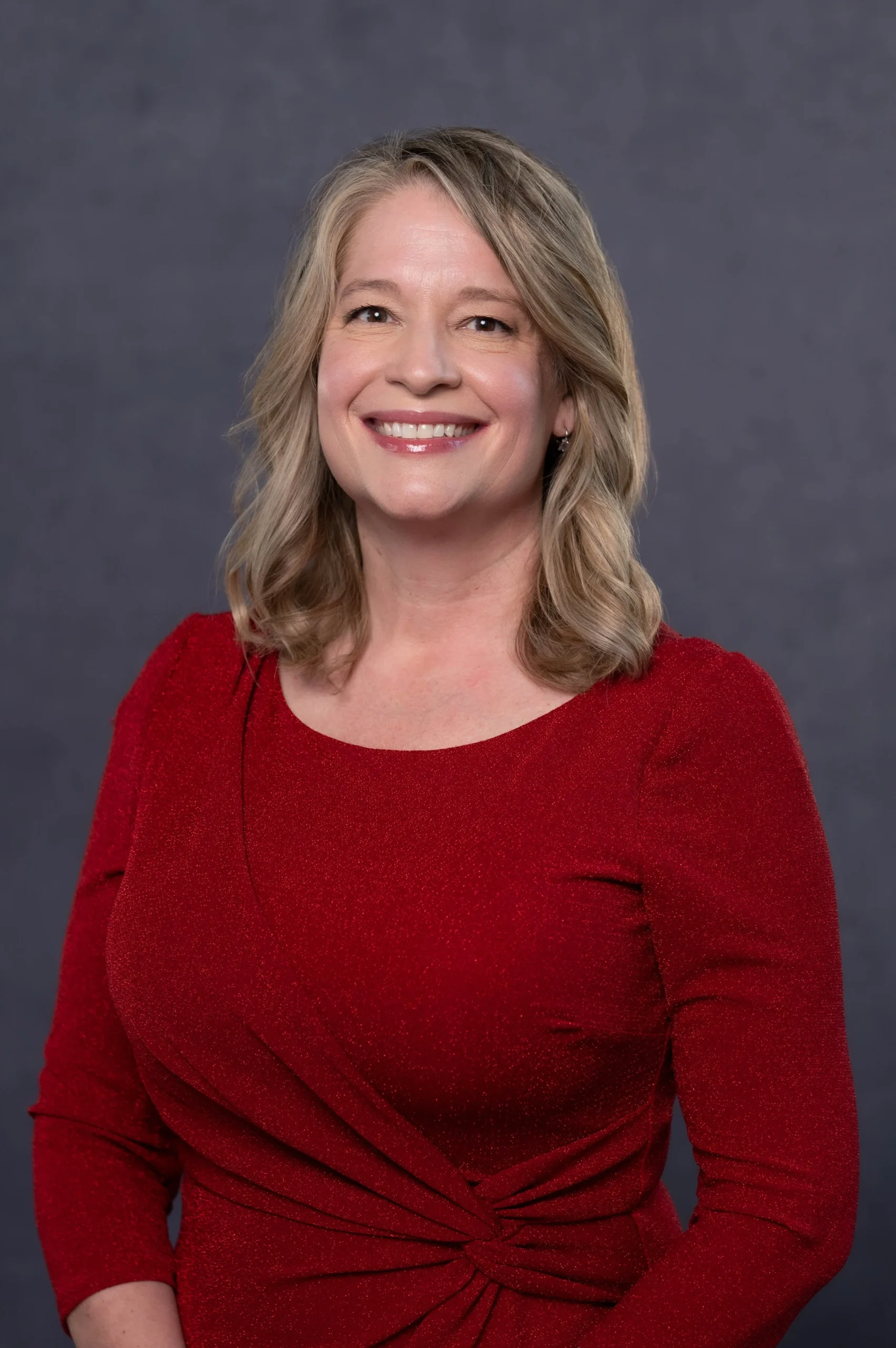 A woman with shoulder-length blonde hair smiling, wearing a red dress with long sleeves and a knot detail at the waist, against a dark gray background.