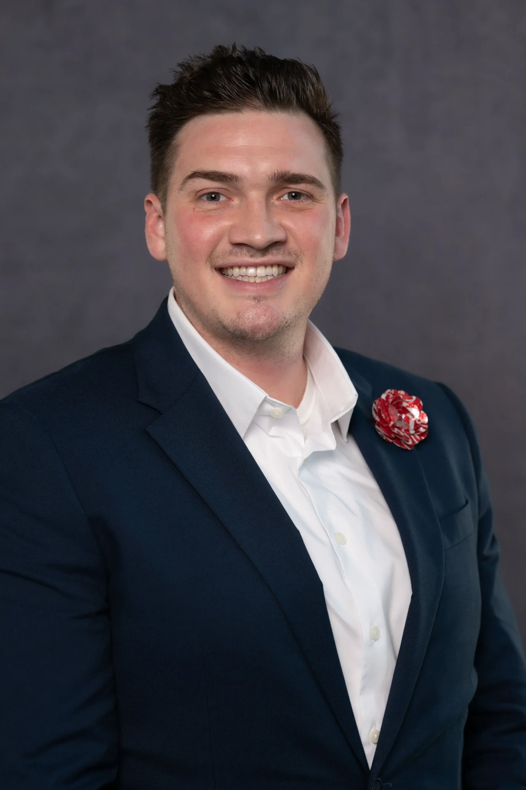 A young man wearing a navy blazer, white dress shirt, and a red patterned lapel pin, smiling against a dark gray background.