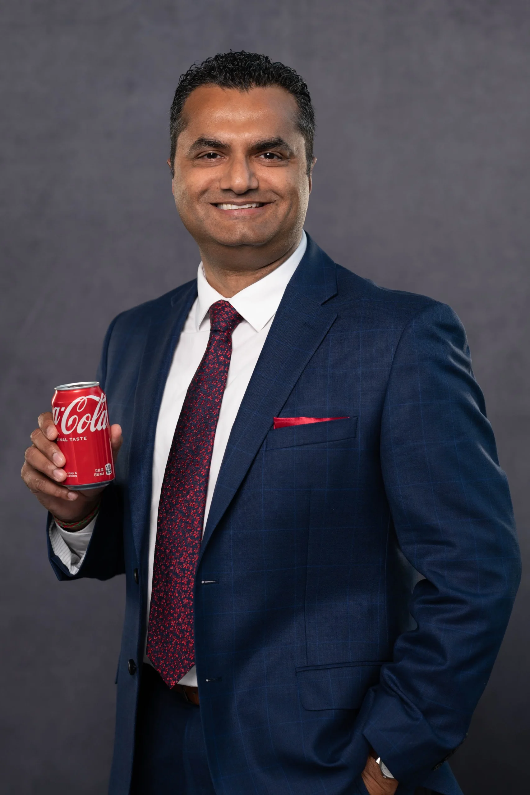 A man in a dark blue suit and red tie holding a can of Coca-Cola, smiling, against a gray background.