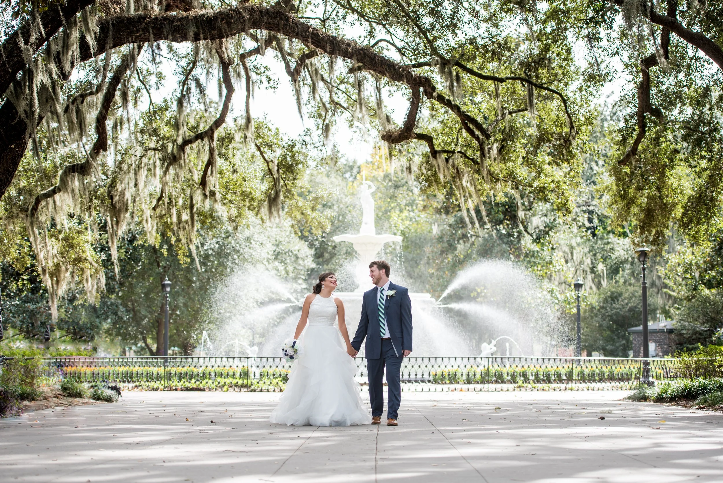 A bride and groom walking hand in hand in a park with a fountain in the background, surrounded by trees with hanging moss.