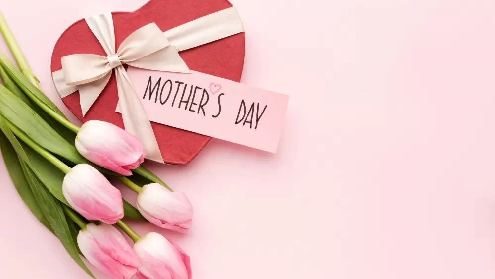 Pink tulips and a red heart-shaped box with a white ribbon, along with a note that says 'Mother's Day' on a pink background.